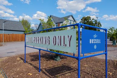Blue-framed sign reading “Hotel Parking Only” marking a hotel parking area by a Tudor-style building, gravel lot, wooden fence and clear blue sky.
