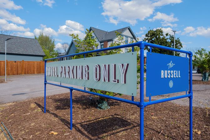 Blue-framed sign reading “Hotel Parking Only” marking a hotel parking area by a Tudor-style building, gravel lot, wooden fence and clear blue sky.