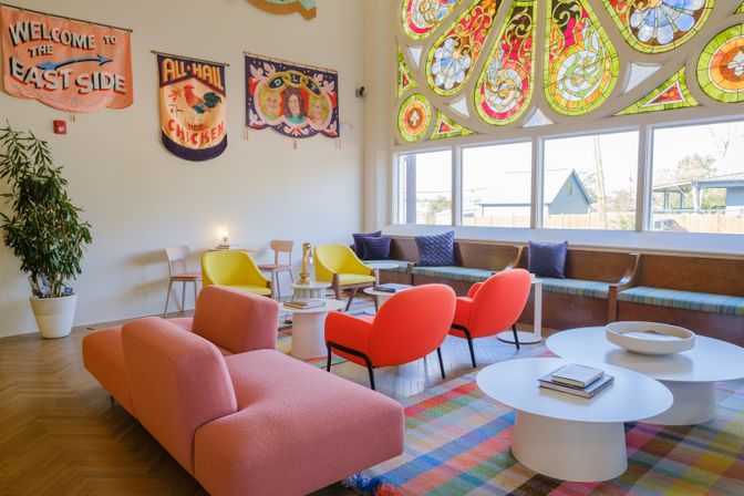 Sunlit eclectic lounge with a large stained-glass window, colorful mid-century seating (pink sofa, red and yellow chairs), plaid rug, and wall banners.