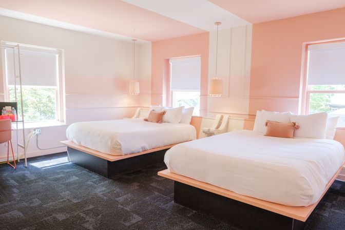 Bright pastel-pink hotel guest room with two white platform beds, tassel throw pillows, hanging pendant lights and large windows with roller shades