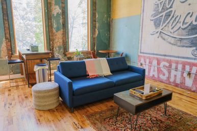 Bright industrial loft lounge with a mid-century blue sofa draped in a pastel throw, hardwood floors and patterned rug, low coffee table with a tray, round knit pouf, exposed brick and peeling plaster walls, tall windows and a small wooden desk with stools.