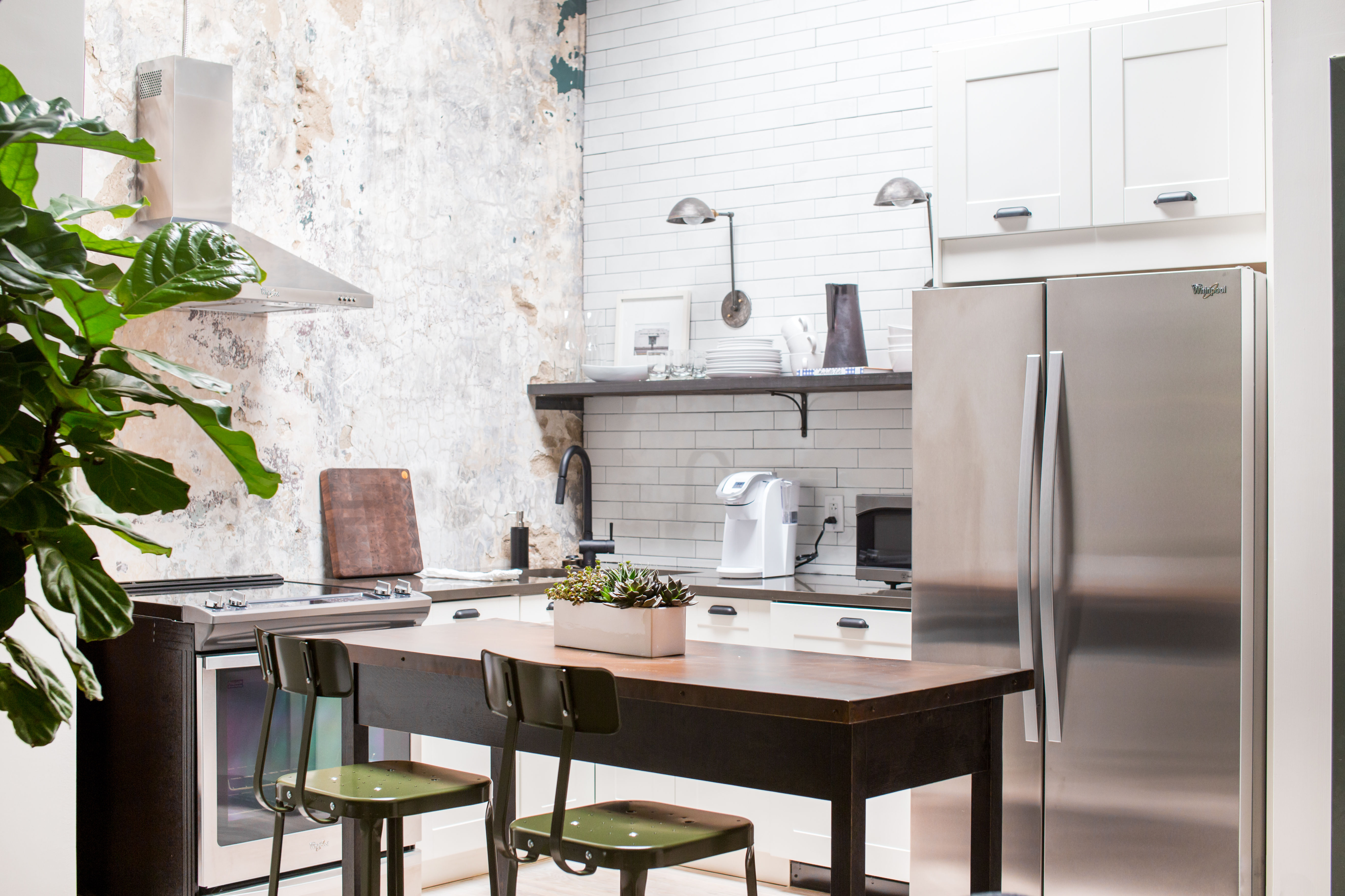 Loft-style modern kitchen with white subway-tile backsplash and distressed plaster wall, stainless-steel refrigerator and range, wooden island with green metal bar stools, succulent centerpiece and large indoor plant.