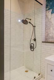 Glass-enclosed shower with white subway tile walls, hexagon tile floor, bronze rain and handheld showerheads, and a textured plaster accent wall.