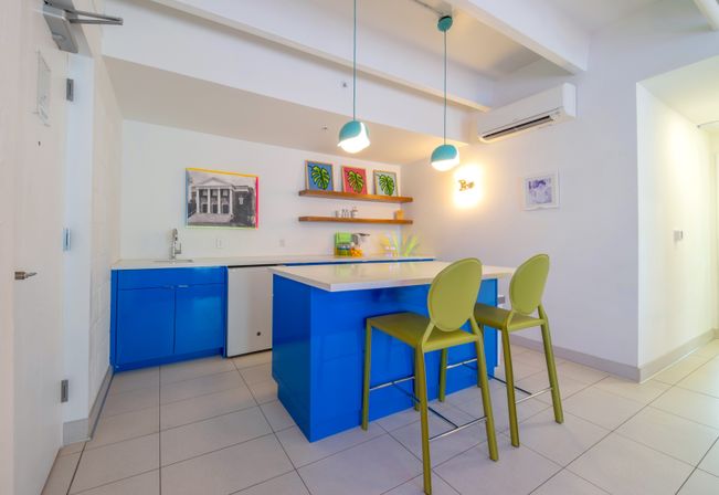 Bright modern kitchenette with glossy blue island and cabinets, white quartz countertop, two lime-green bar stools, teal pendant lights, floating wooden shelves with tropical leaf prints, small sink and mini-fridge on tiled floor.
