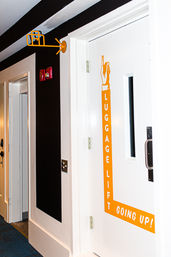 Hotel corridor with a white door labeled LUGGAGE LIFT — GOING UP! in bright orange, matching arrow and suitcase icon overhead, dark accent wall, nearby guest room doors and blue carpeted floor.