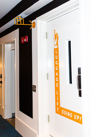 Hotel corridor with a white door labeled LUGGAGE LIFT — GOING UP! in bright orange, matching arrow and suitcase icon overhead, dark accent wall, nearby guest room doors and blue carpeted floor.