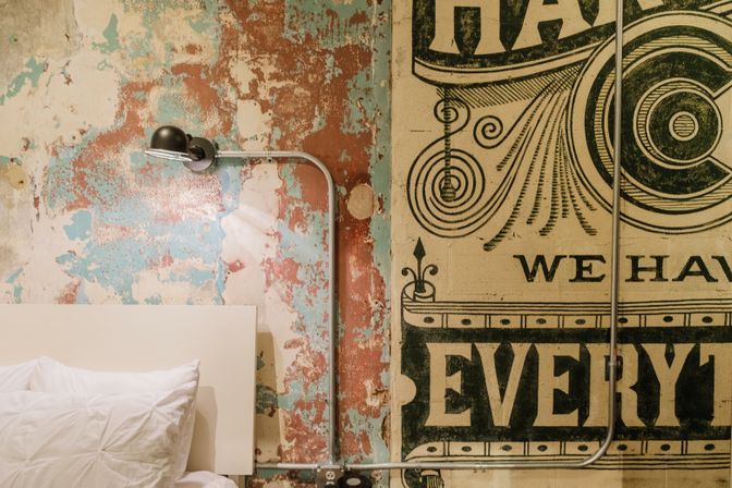 Industrial-style bedroom close-up: weathered turquoise and rust peeling paint beside a vintage black graphic mural, exposed metal conduit and wall lamp above a white headboard and pillow.