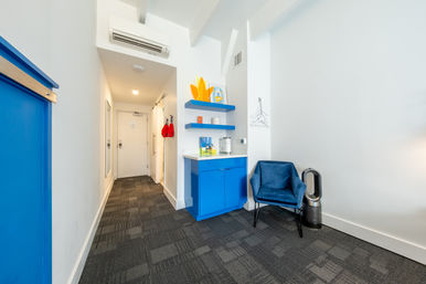 Bright modern hotel room entry with bold blue accents: compact kitchenette and floating shelves, velvet navy chair next to an air purifier, carpeted hallway leading to a white door and wall hooks