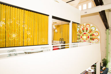 Bright modern mezzanine loft with mustard-yellow velvet curtains, glass-walled meeting room, white railing and a colorful stained-glass rose window.