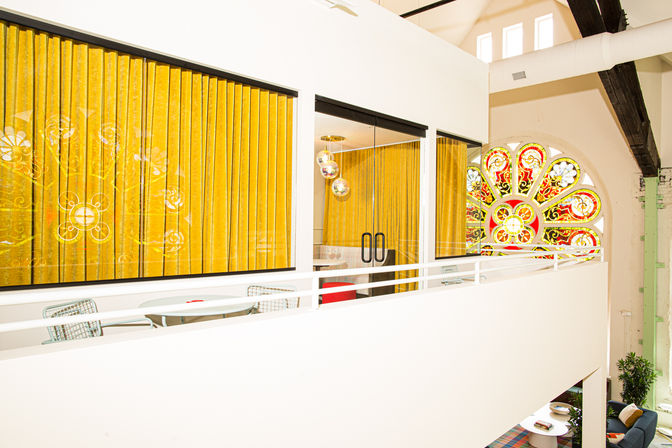 Bright modern mezzanine loft with mustard-yellow velvet curtains, glass-walled meeting room, white railing and a colorful stained-glass rose window.