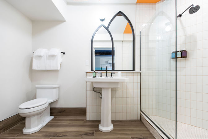 Bright modern bathroom interior with white pedestal sink, black arched mirror, glass walk-in shower with white tiles, toilet, folded towels and wood-look tile floor