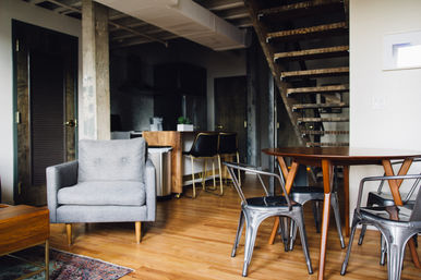Cozy modern loft living area with a gray armchair, round wooden dining table, industrial metal chairs, kitchen island and open wooden staircase on warm hardwood floors.