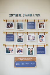 Hotel lobby charity wall display in Nashville with brass rods and leather straps holding photo panels and blue info cards under bold lettering “STAY HERE. CHANGE LIVES.” showcasing local volunteers and partner organizations.