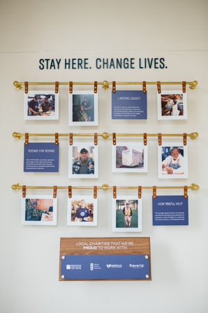 Hotel lobby charity wall display in Nashville with brass rods and leather straps holding photo panels and blue info cards under bold lettering “STAY HERE. CHANGE LIVES.” showcasing local volunteers and partner organizations.