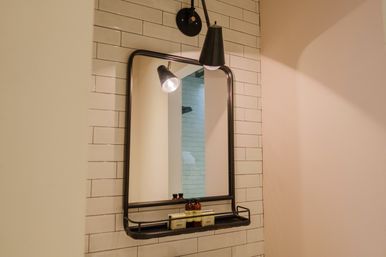 Contemporary bathroom wall with a black-framed mirror and built-in shelf holding small toiletry bottles and soap against white subway tile, lit by a black sconce.