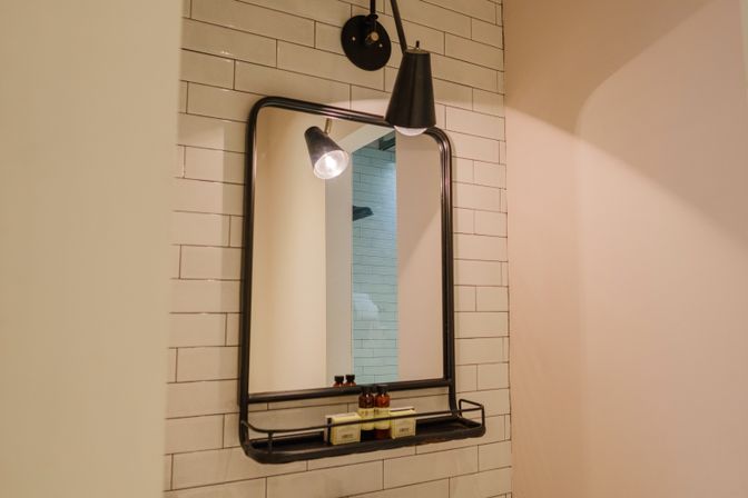 Contemporary bathroom wall with a black-framed mirror and built-in shelf holding small toiletry bottles and soap against white subway tile, lit by a black sconce.