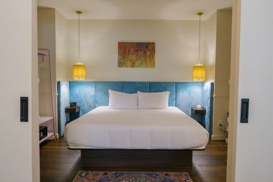 Cozy modern hotel bedroom with king bed and crisp white linens, blue upholstered headboard, two yellow pendant lamps, colorful abstract wall art, wooden floor and patterned rug