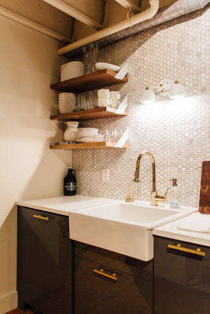 Chic modern kitchen nook with white apron-front sink, brass faucet, hexagon tile backsplash and wooden floating shelves holding white dishes and glassware.