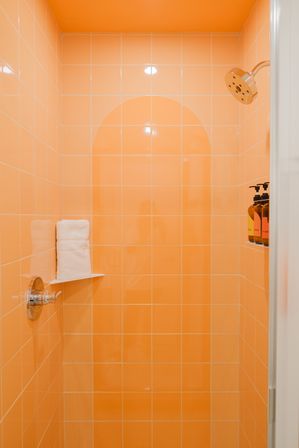 Bright orange tiled shower stall with arched tile accent, chrome showerhead and control, folded white towels on a corner shelf and amber pump bottles in a recessed niche