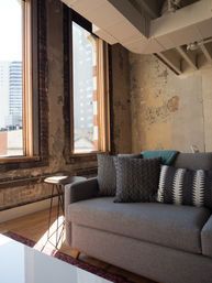 Sunlit urban loft living room with tall wood-framed windows, distressed exposed brick and peeling plaster walls, a gray sofa with textured and striped pillows, small round metal side table and hardwood floor with city buildings visible outside.