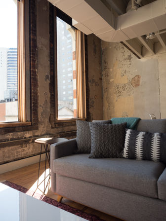 Sunlit urban loft living room with tall wood-framed windows, distressed exposed brick and peeling plaster walls, a gray sofa with textured and striped pillows, small round metal side table and hardwood floor with city buildings visible outside.