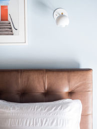 Close-up of a modern bedroom: brown tufted leather headboard against a pale blue wall, crisp white pillow, white brass-accent wall sconce and framed photo of stairs with an orange door.