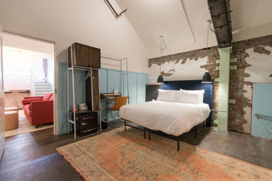 Urban industrial loft bedroom with a crisp white bed and navy headboard against exposed brick, black pendant lights, mint-painted support beam, vintage faded rug, small desk, and an open doorway revealing a red sofa lounge.