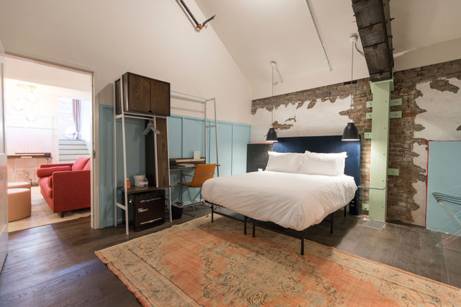 Urban industrial loft bedroom with a crisp white bed and navy headboard against exposed brick, black pendant lights, mint-painted support beam, vintage faded rug, small desk, and an open doorway revealing a red sofa lounge.