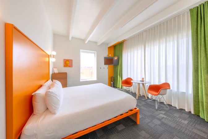 Bright modern hotel room with a king bed and bold orange headboard, crisp white linens, green floor-to-ceiling curtains, two orange lounge chairs with a small round table by a large window, and a wall-mounted TV — playful orange and green accents.
