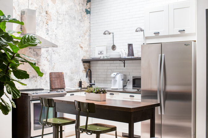 Bright modern kitchen with stainless steel refrigerator, subway tile backsplash, wooden island with green metal bar stools, potted succulents and a large leafy plant.
