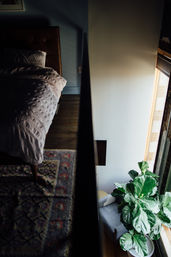 Top-down view of a cozy urban loft bedroom: rumpled bed and patterned rug beside a dark railing overlooking a sunlit window nook with a gray chair and large fiddle-leaf fig plant.