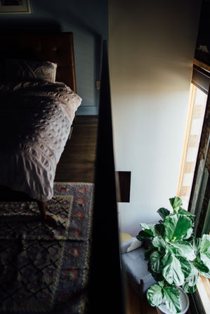 Top-down view of a cozy urban loft bedroom: rumpled bed and patterned rug beside a dark railing overlooking a sunlit window nook with a gray chair and large fiddle-leaf fig plant.