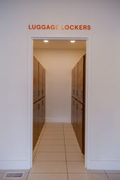 Entrance to luggage lockers — narrow tiled corridor lined with wooden storage lockers on both sides under an orange "LUGGAGE LOCKERS" sign.