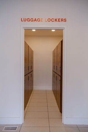 Entrance to luggage lockers — narrow tiled corridor lined with wooden storage lockers on both sides under an orange "LUGGAGE LOCKERS" sign.