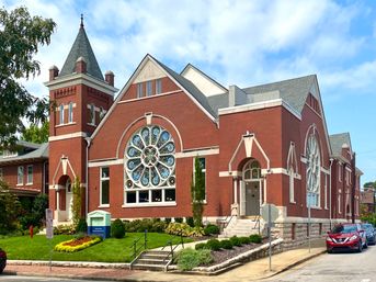 Red-brick historic church on a neighborhood corner with a turreted tower, ornate circular stained-glass rose windows, arched stone entrances, front steps and manicured lawn under a bright blue sky.
