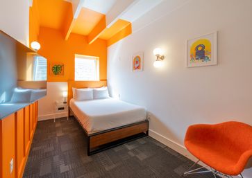 Cozy urban boutique hotel room with bright orange accent wall and ceiling beams, queen bed with white linens, framed wall art, wall sconces, flat-screen TV, and an orange mid-century chair.