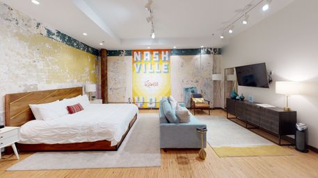 Bright Nashville loft-style bedroom with distressed plaster walls and a colorful mural reading NASHVILLE, king wood bed with white linens, light blue sofa, armchair, wall-mounted TV and modern lamps on hardwood floors.