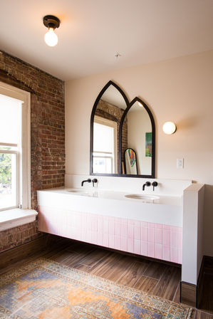 Loft-style bathroom with floating double vanity with pink tile front, two black arched mirrors, matte black wall faucets, exposed brick wall, round wall sconce, wood-look floors and patterned rug by a sunlit window.