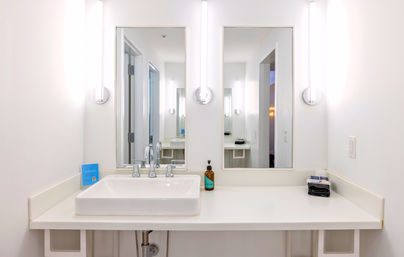 Bright minimal white bathroom vanity with rectangular vessel sink, chrome faucet, twin mirrors and tall illuminated wall sconces, amber soap bottle and folded dark towels on the countertop.