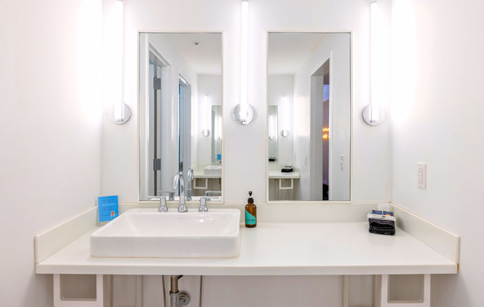 Bright minimal white bathroom vanity with rectangular vessel sink, chrome faucet, twin mirrors and tall illuminated wall sconces, amber soap bottle and folded dark towels on the countertop.