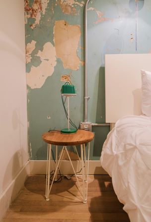 Cozy bedroom corner: wooden nightstand with a green vintage lamp, peeling teal plaster wall and exposed metal conduit beside a white bed