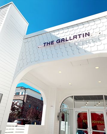 Bright blue-sky view of a white coastal-style building entrance with scalloped shingle facade, purple-letter sign, arched canopy and glass double doors.