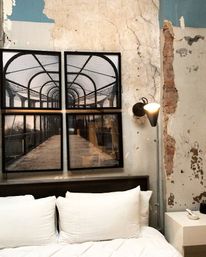 Industrial urban loft bedroom with white pillows and bedding, distressed plaster and exposed brick wall, four-panel framed photo of a caged pedestrian bridge, cone wall sconce and white nightstand.