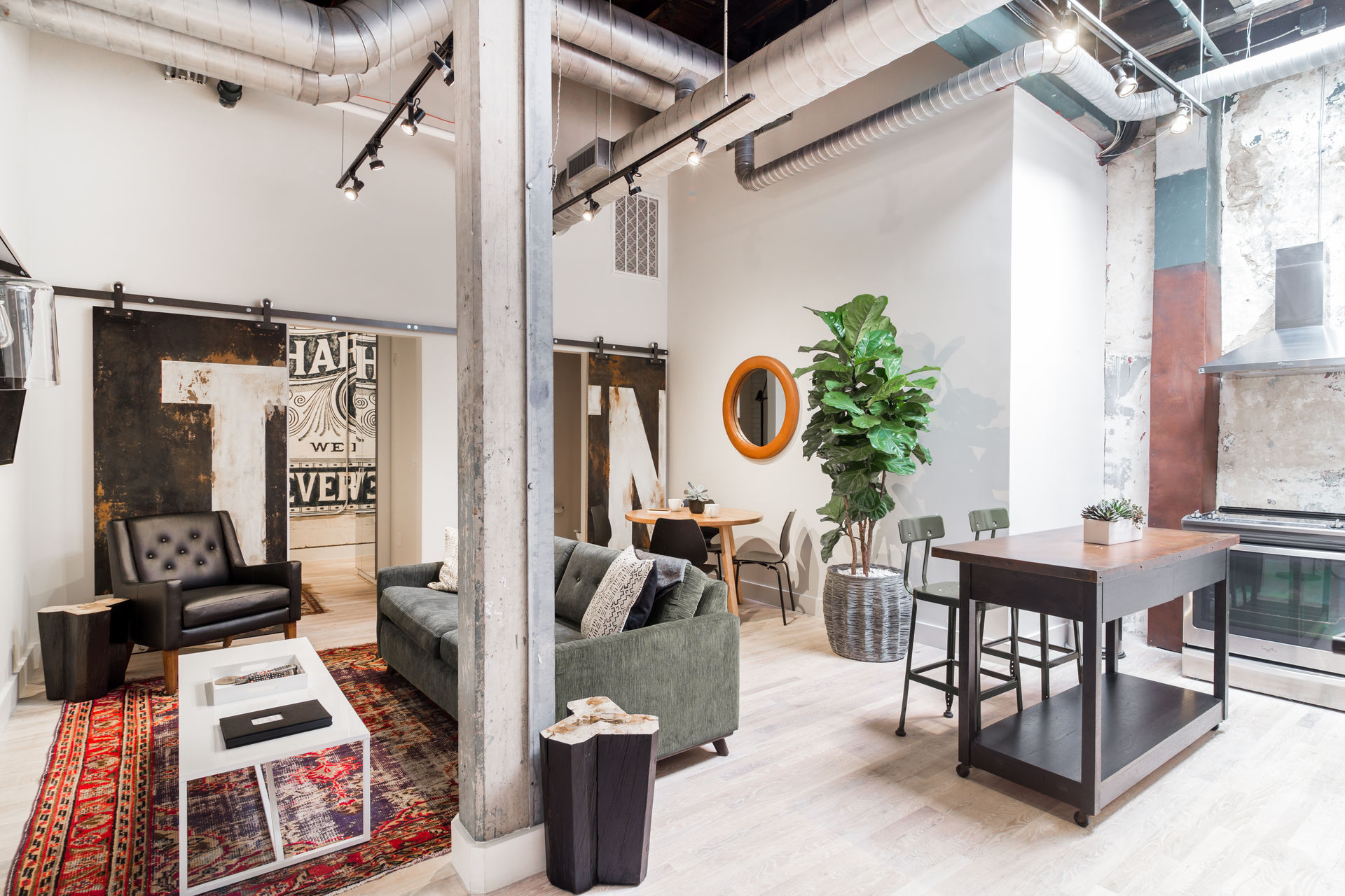Sunlit industrial loft interior with exposed ductwork, green sofa and black leather armchair on a red patterned rug, round orange mirror, fiddle‑leaf fig, and a wooden kitchen island with metal stools.