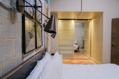Loft-style industrial bedroom with exposed concrete block wall, black adjustable wall lamp and framed art above a leather headboard, crisp white pillows and bedding, frosted sliding closet doors, warm recessed lighting and a patterned rug.