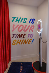 Playful indoor selfie/photo booth alcove with illuminated ring light, red velvet curtain, blue floor mat and a colorful wall mural that reads “This is your time to shine!”