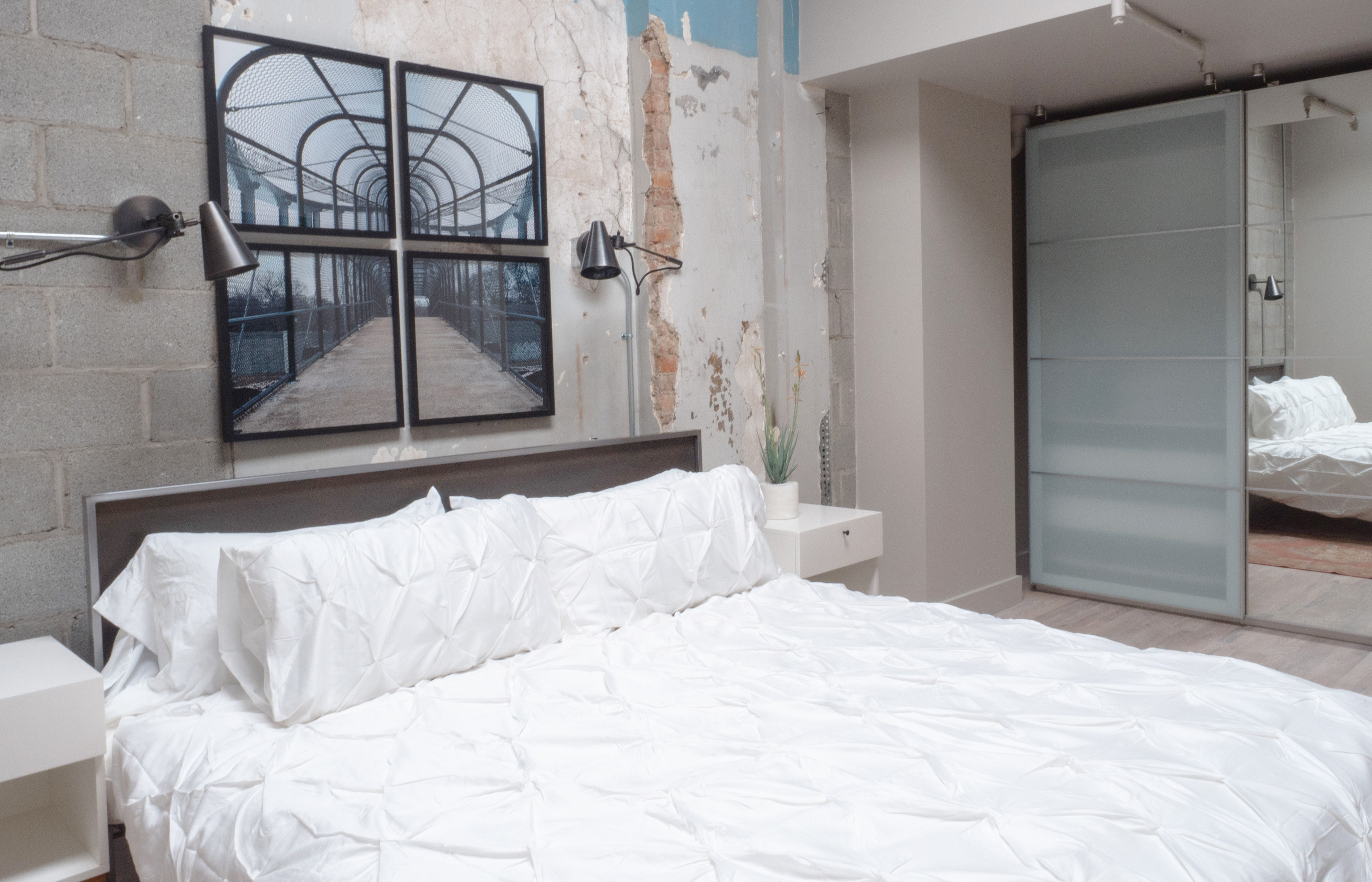 Modern industrial loft bedroom with exposed concrete and peeling plaster wall, white textured duvet and pillows, black swing-arm lamps, four-panel walkway artwork and frosted sliding closet doors.