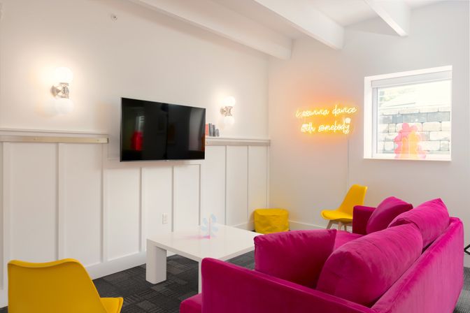 Bright modern lounge with fuchsia velvet sofa, yellow accent chairs, white paneled walls, wall-mounted TV and playful neon sign reading i wanna dance with somebody by a window