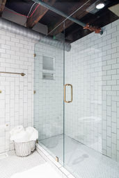 Bright modern bathroom with glass-enclosed white subway-tile shower, brass door handle, penny-tile floor, exposed wooden beams and ductwork, and a wire basket of rolled white towels.