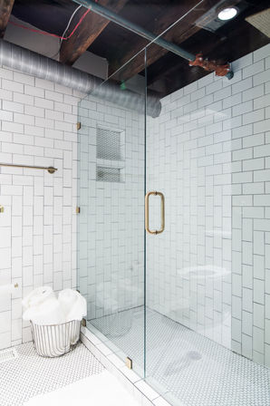 Bright modern bathroom with glass-enclosed white subway-tile shower, brass door handle, penny-tile floor, exposed wooden beams and ductwork, and a wire basket of rolled white towels.